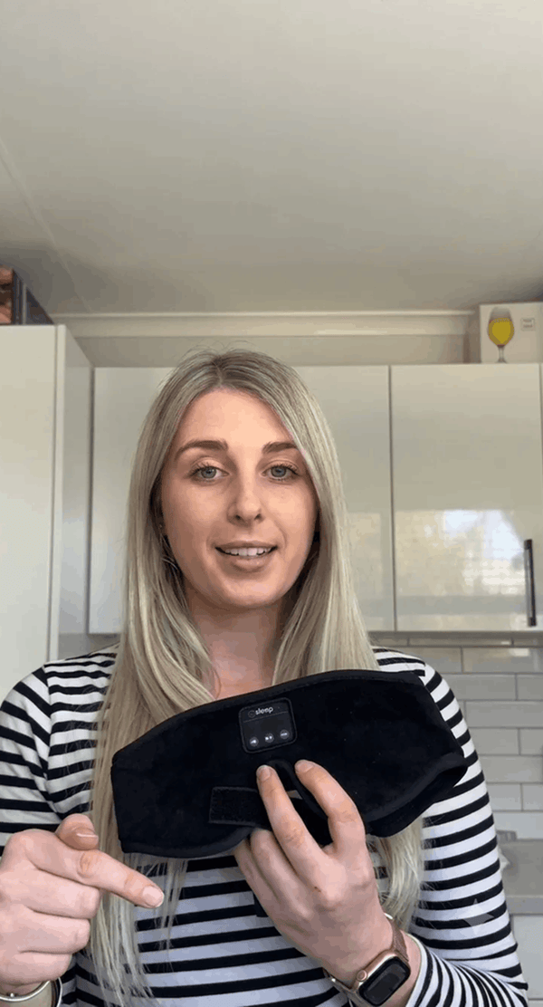 A woman with blonde hair wearing a black and white striped shirt holds up Dreamy Sounds Sleep Mask | Bluetooth Sleep Mask in a kitchen setting.