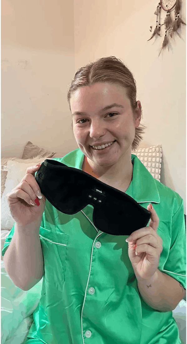 A woman in a green satin pajama shirt holds up Dreamy Sounds Sleep Mask | Bluetooth Sleep Mask in front of her face, smiling in a bedroom setting.