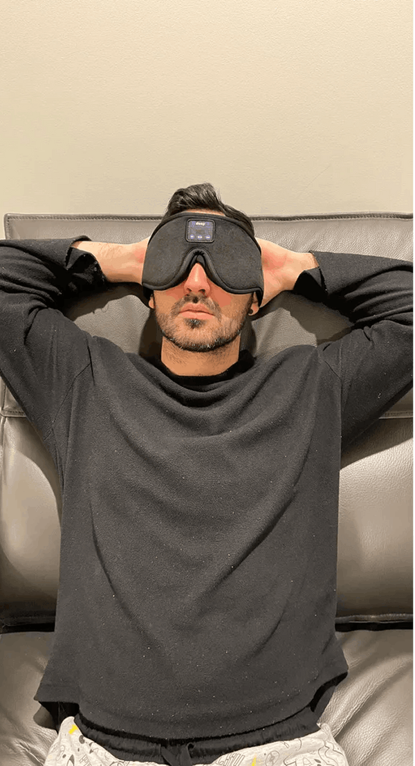 A man reclining on a gray leather couch, wearing Dreamy Sounds Sleep Mask | Bluetooth Sleep Mask. He is dressed in a dark gray long-sleeve shirt and light-colored pants.